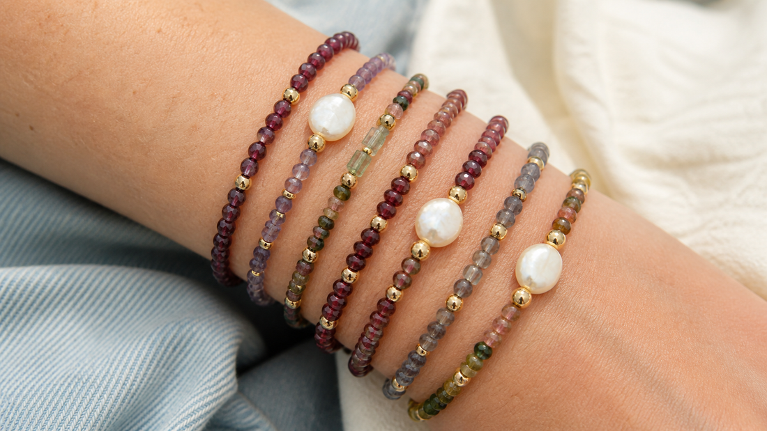 Seven stacked crystal bracelets in wine red, moss green, smoky purple, and pearl tones with gold spacers