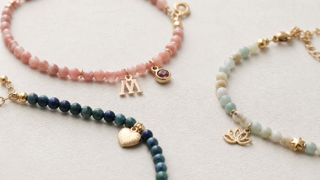 Three personalized gemstone bracelets with initial, heart, and lotus charms on ivory fabric